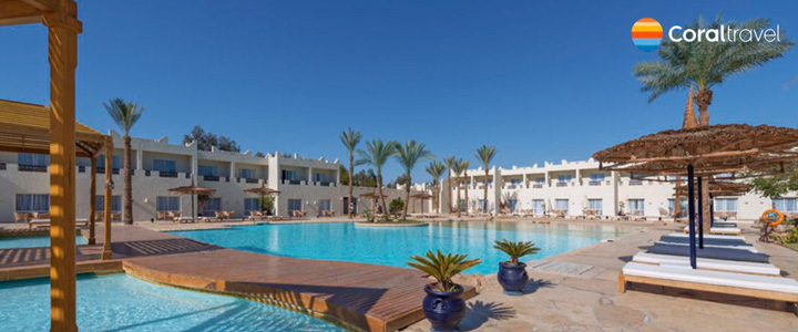 Vom Hotel eröffnet sich der Blick über die weitläufige Poollandschaft und den grünen Park bis hin zum Strand mit Sonnenliegen, dahinter liegt das Rote Meer im warmen Licht des Sonnenuntergangs.